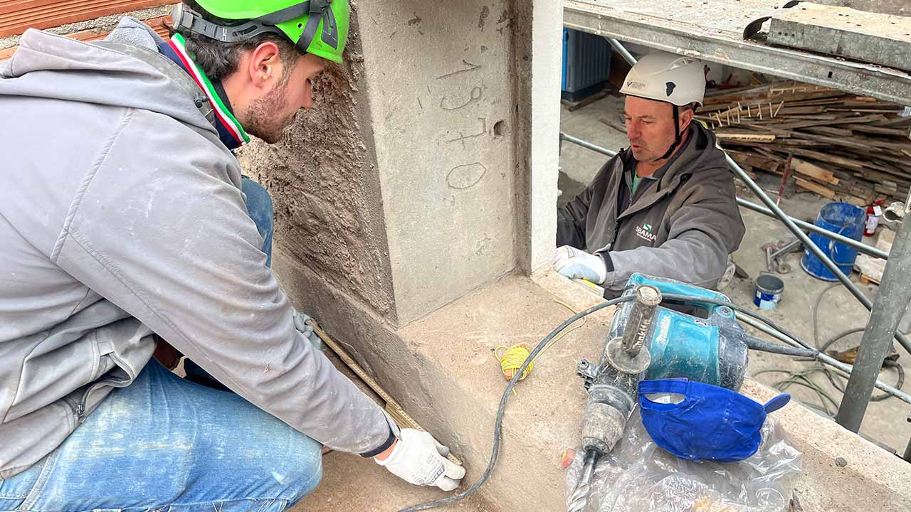 Due uomini misurano una parete di una casa, uno dall'esterno e l'altro dall'interno, con attorno attrezzi di restaurazione dell'immobile